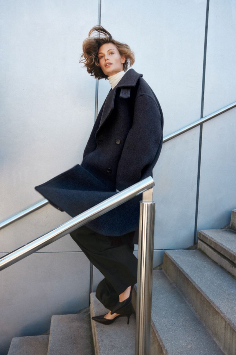 Fashionable woman in coat on stairs: Elegant outerwear, black pants and pumps. Street style, urban setting, hair blowing in wind.