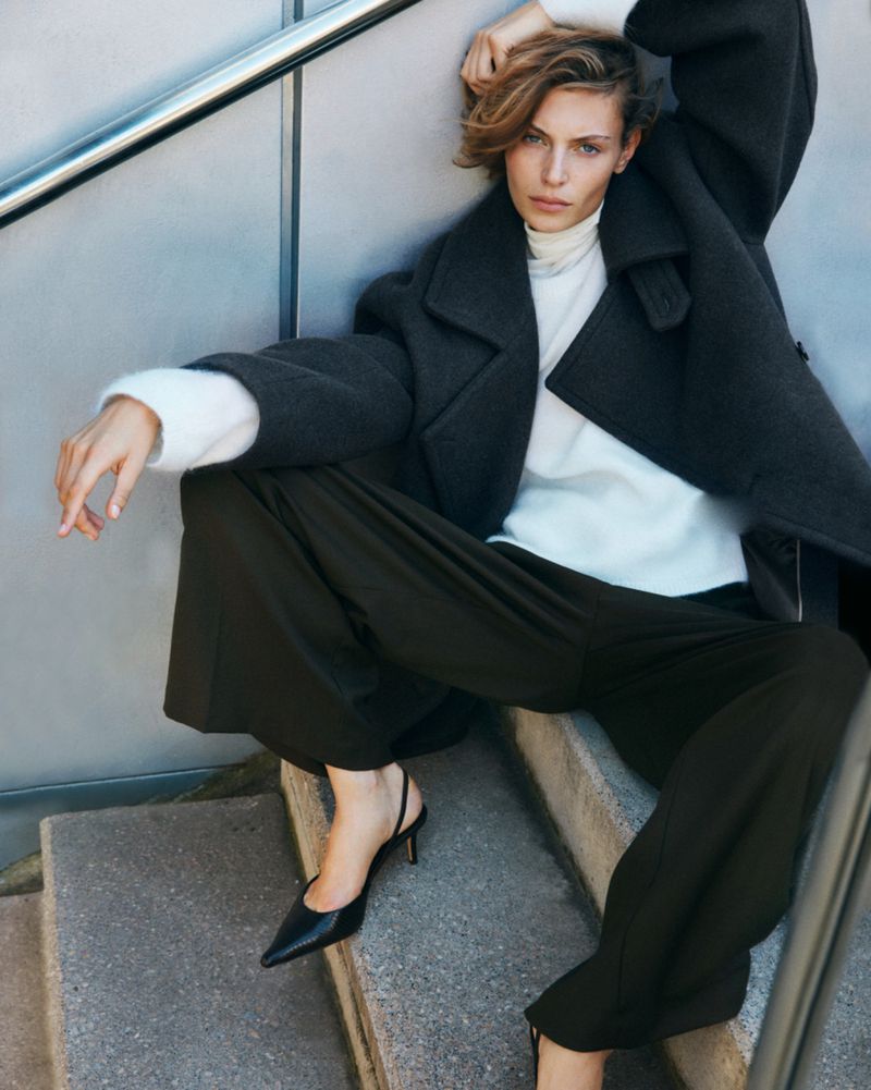 A fair-skinned woman with short blonde hair poses on concrete steps wearing a dark overcoat, cream turtleneck, black trousers, and black slingbacks.