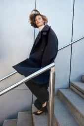 Model wearing a dark wool coat and black trousers standing on concrete stairs beside a metallic handrail.