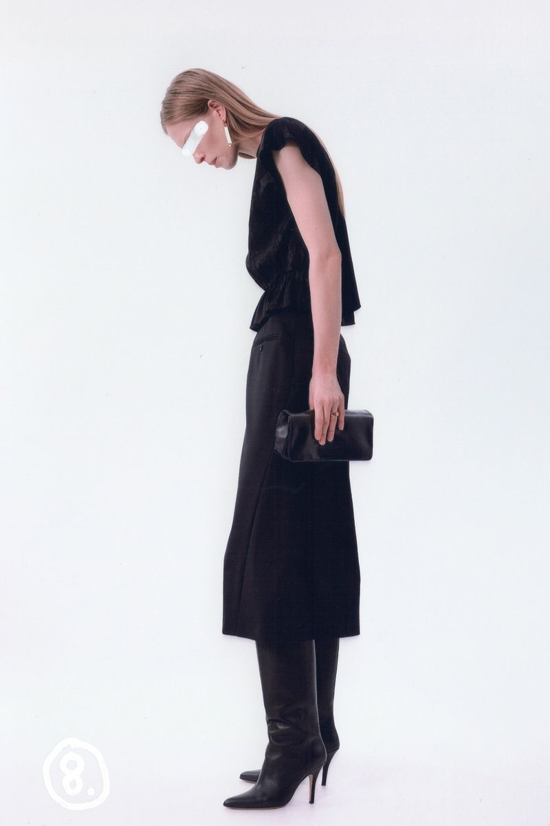 Fashion model in a studio wearing a dark top, skirt, and boots while holding a clutch. The background is white, and the lighting is soft.
