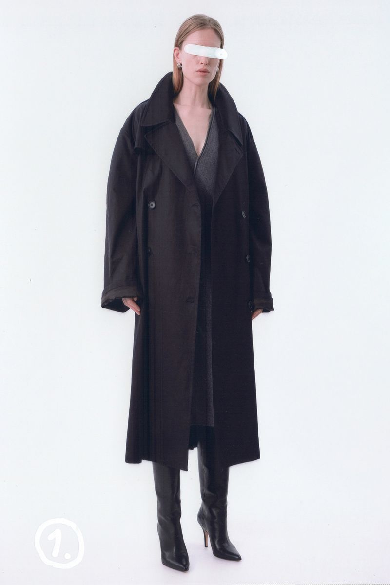 Fashion editorial shot of a blonde female model in a long, black trench coat and high-heeled boots against a bright white studio backdrop.