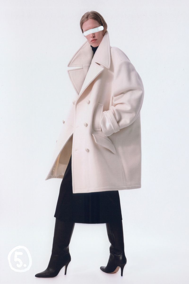 A fashion shot of a model wearing a double-breasted coat with black boots, posing against a white background in a studio setting.