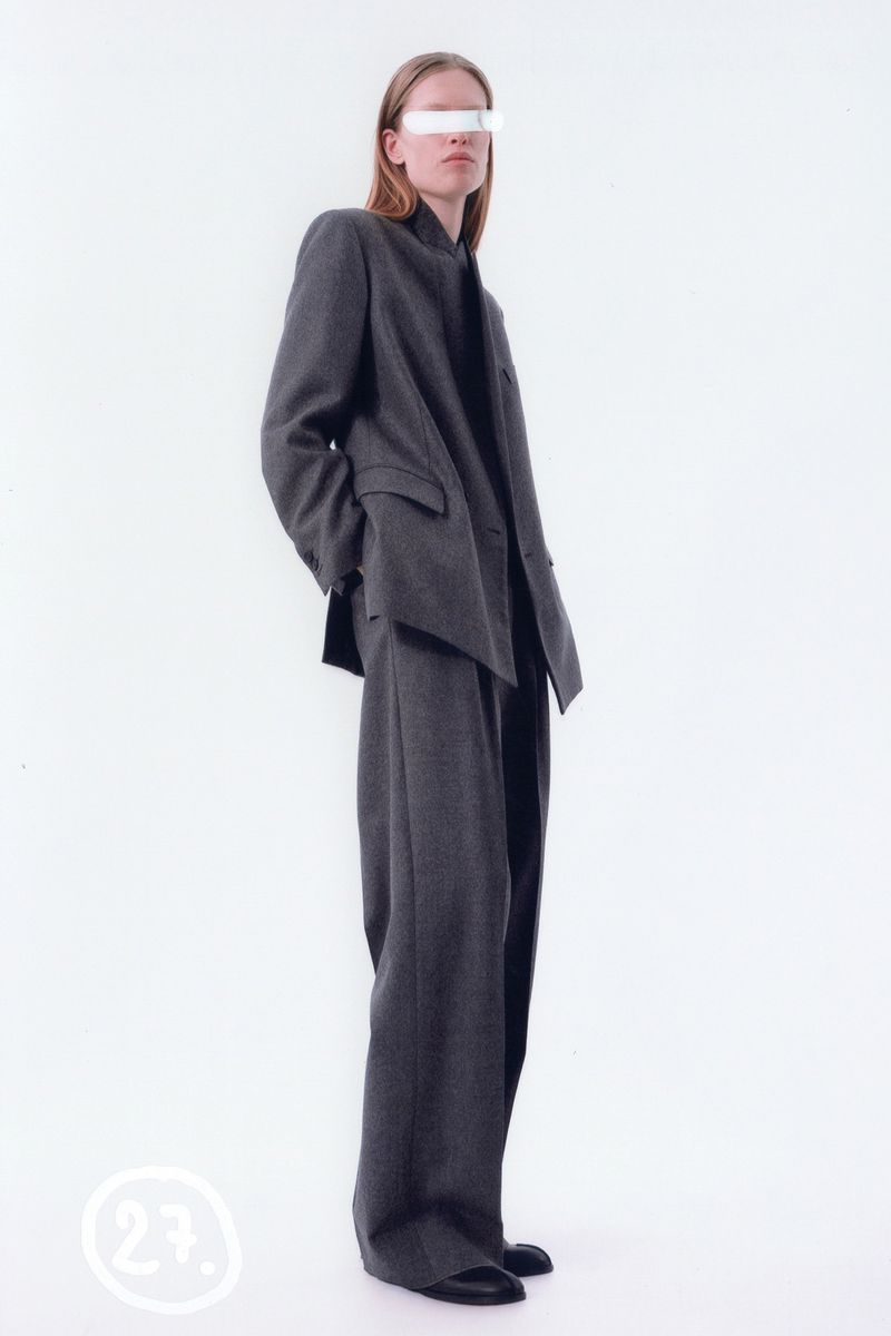 A tall fashion model poses in a grey suit in front of a white background in a studio with even lighting wearing a white censor bar.