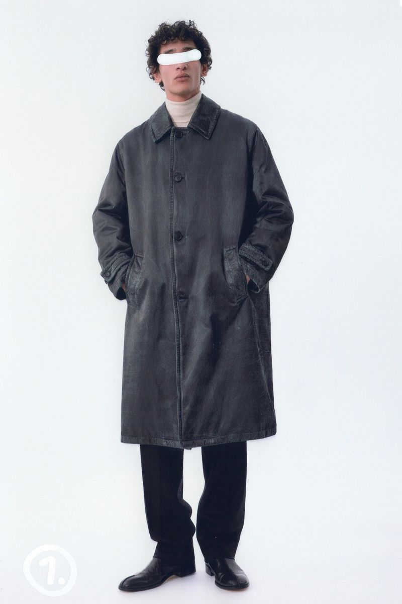 Full shot of a man in an overcoat, dress pants, and dress shoes, posing in a bright white studio setting with a minimalist aesthetic.