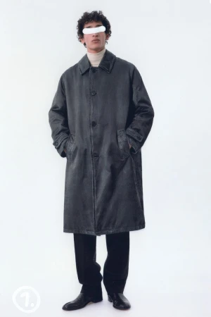 Full shot of a man in an overcoat, dress pants, and dress shoes, posing in a bright white studio setting with a minimalist aesthetic.