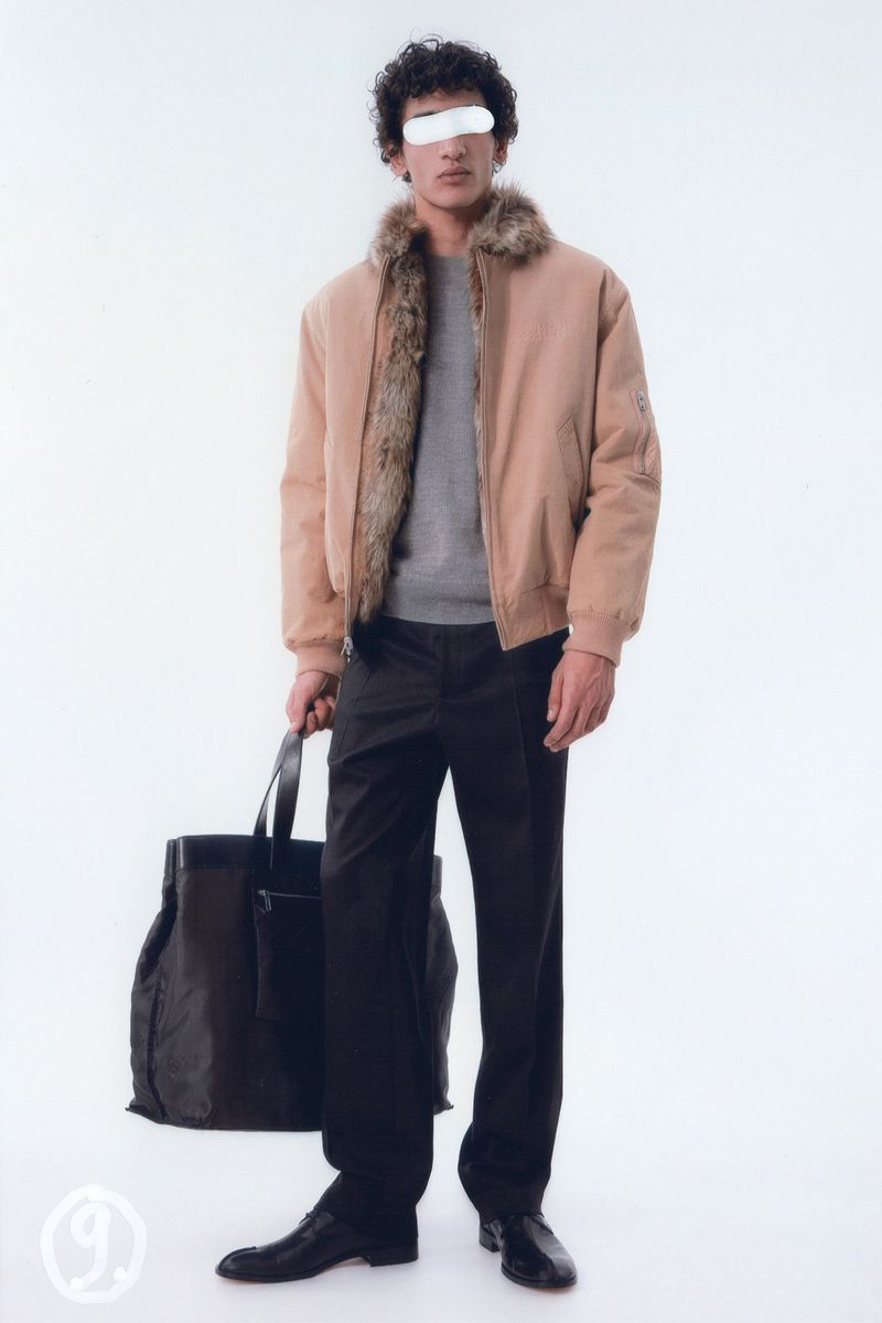 A man in a beige bomber jacket with a fur collar, grey shirt, black pants, and black shoes holds a black tote bag in a studio shot.