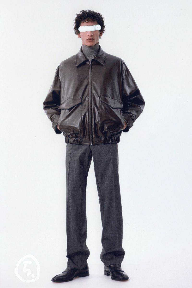 Fashionable adult male posing in a brown leather jacket, gray turtleneck, trousers, and black loafers against a white background.