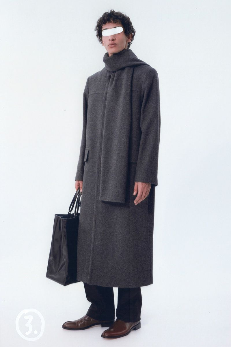 An adult male fashion model wears a long gray coat, black pants, and brown shoes while holding a black bag against a white background.