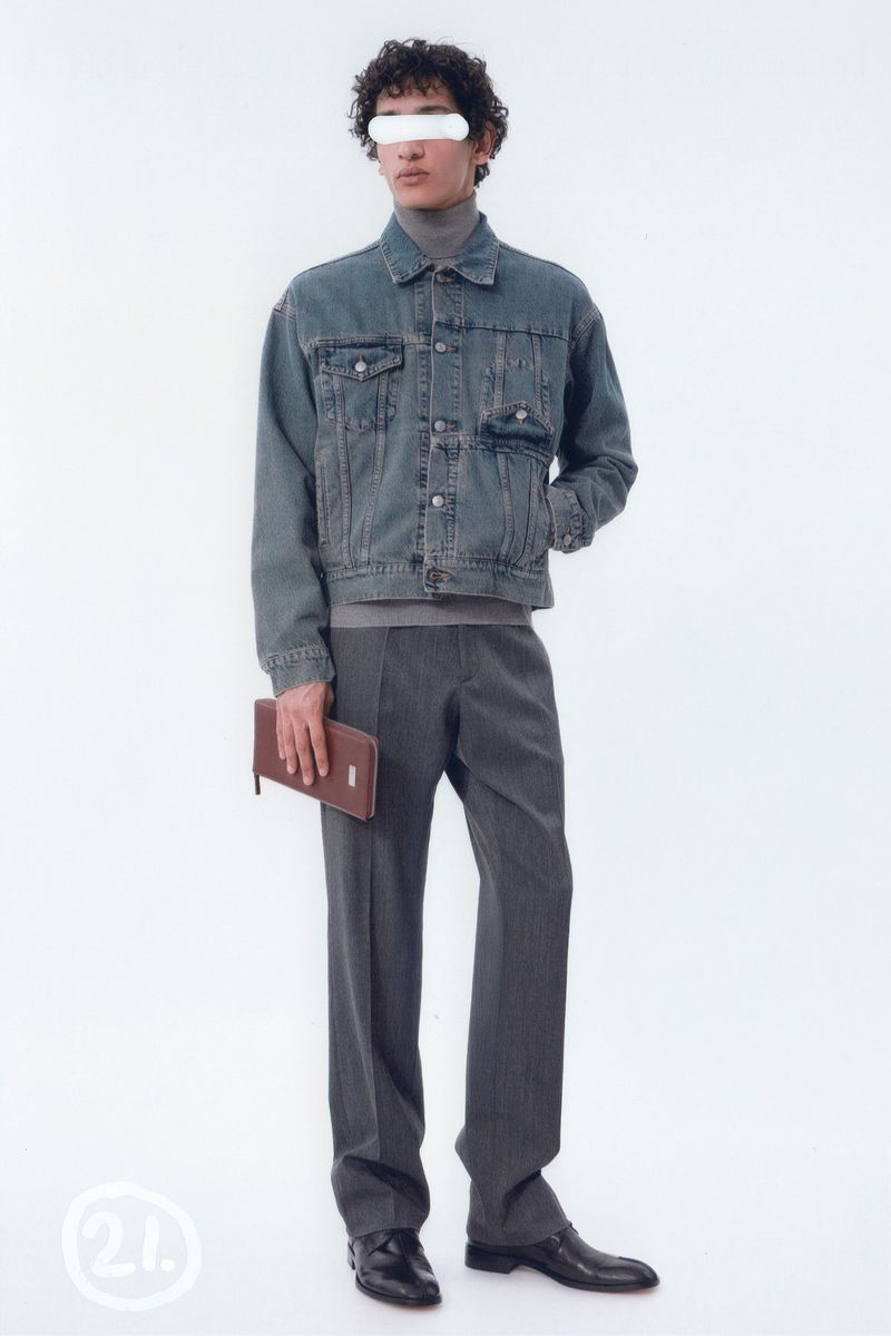 Full-length studio shot of a man in a denim jacket, grey trousers, and black shoes, holding a brown wallet against a white background.