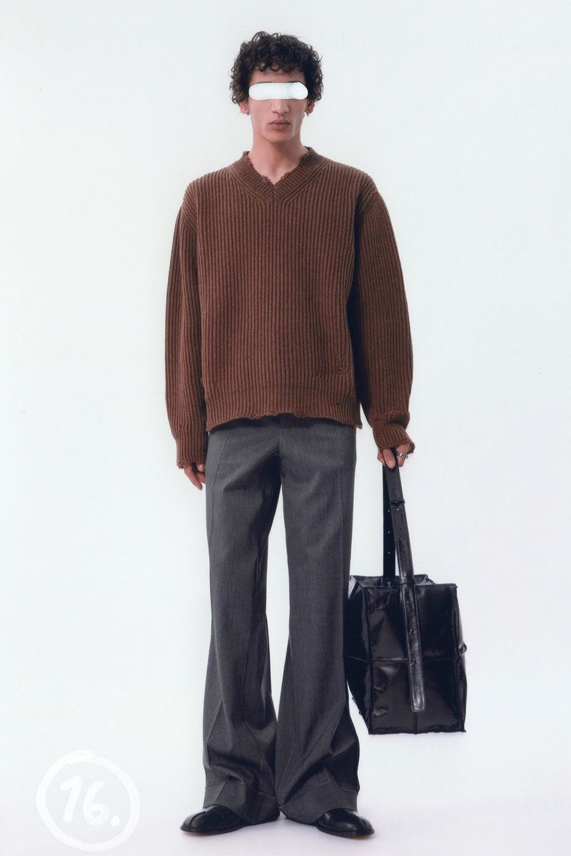 Full-length studio shot of a fashion model wearing a brown v-neck sweater, grey slacks, and black shoes and holding a black tote bag.
