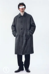 Model wearing a dark grey coat and black trousers standing against a white studio background.