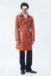 Model wearing a rust-colored leather trench coat and black trousers against a plain white studio background.