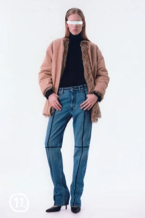Model wearing a tan jacket over a black turtleneck and blue denim jeans against a plain white background