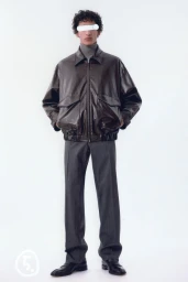 Model wearing a dark leather bomber jacket and grey trousers against a white seamless background
