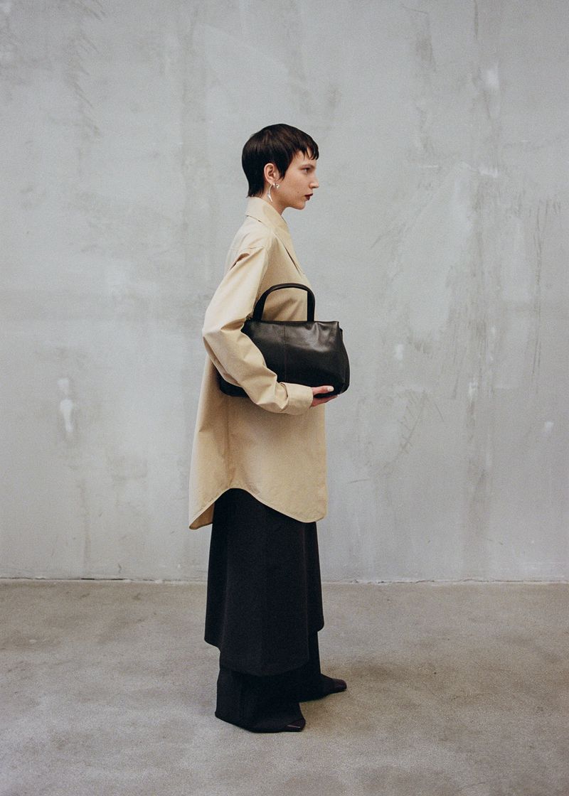 Fashion shot of a woman with short hair wearing a light beige oversized shirt, dark pants, and holding a black leather handbag.