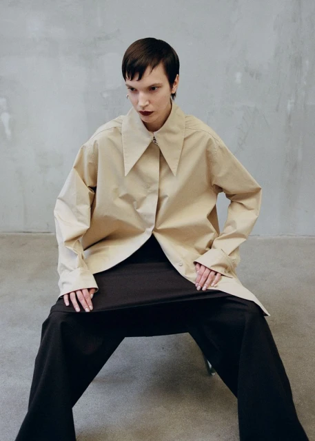 A woman with short dark hair models a beige oversized shirt and black wide-leg pants against a neutral grey background in a studio shot.