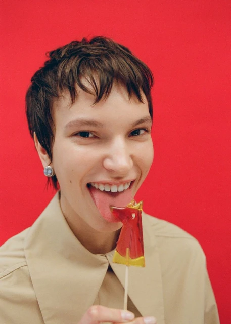 Close-up portrait of a young adult female with short brown hair, sticking out her tongue with a lollipop against a red background.