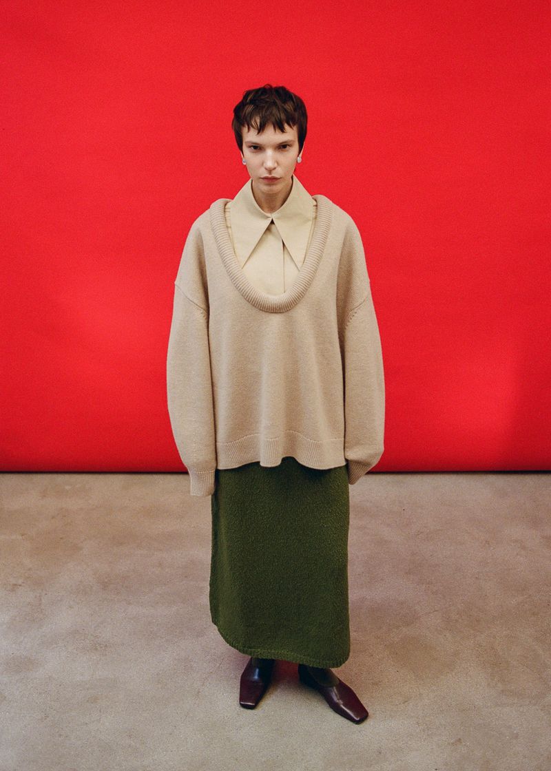 Model wearing a cream scoop-neck sweater and green skirt against a red backdrop