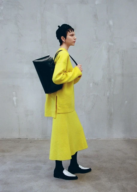Fashion shot of a woman wearing a yellow sweater and skirt carrying a black bag over her shoulder against a textured grey background.