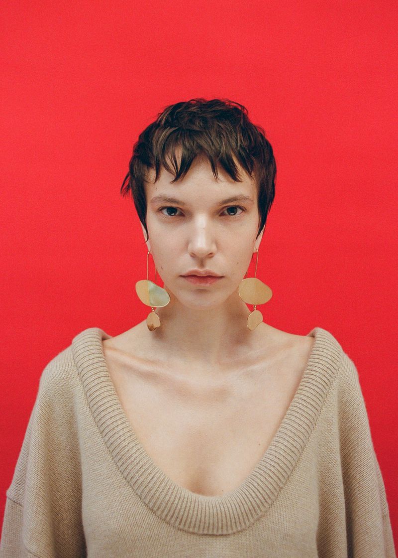 Close-up portrait of a woman with short dark hair wearing large abstract gold earrings and a beige knit sweater against a vibrant red backdrop.