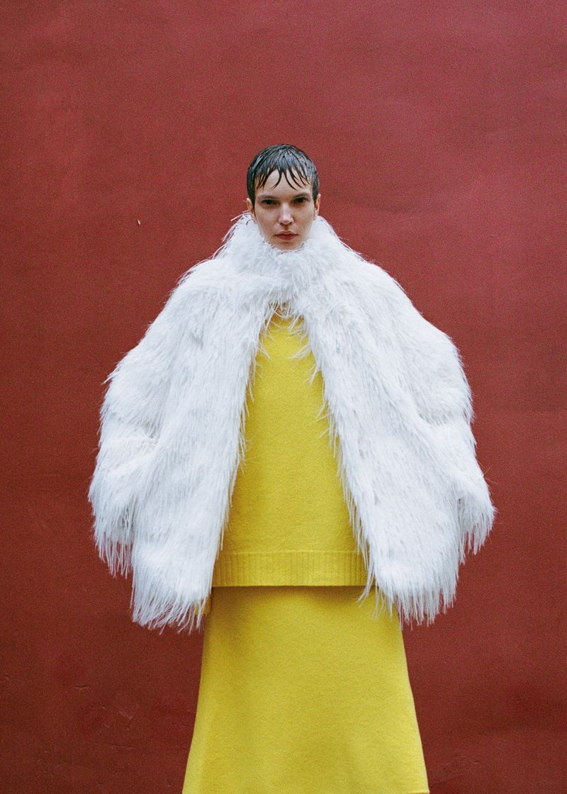 An adult female model poses in a fluffy white coat over a yellow sweater and skirt against a plain red background in a fashion shot.