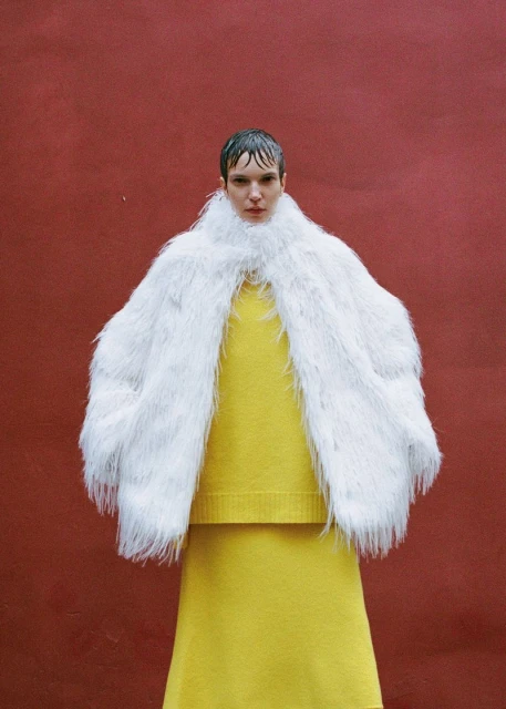 An adult female model poses in a fluffy white coat over a yellow sweater and skirt against a plain red background in a fashion shot.