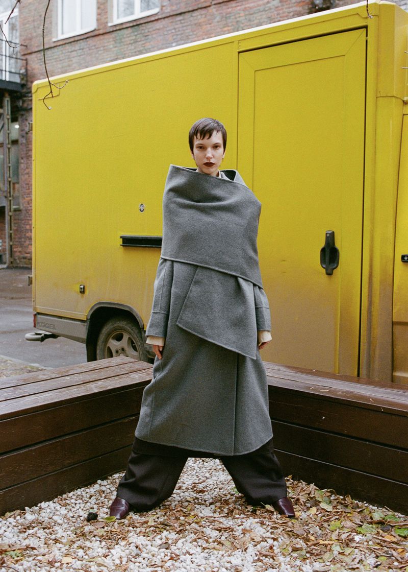 Fashionable woman in a grey coat poses on a gravel surface with a yellow truck and brick building as a backdrop in an outdoor setting.