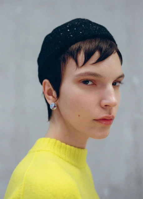 Close-up portrait of a young woman with short dark hair wearing a black knit hat and yellow sweater against a grey background.