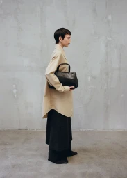 Model in profile wearing an oversized beige button-down shirt and dark skirt holding a black leather handbag