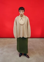 Model wearing a cream scoop-neck sweater and green skirt against a red backdrop