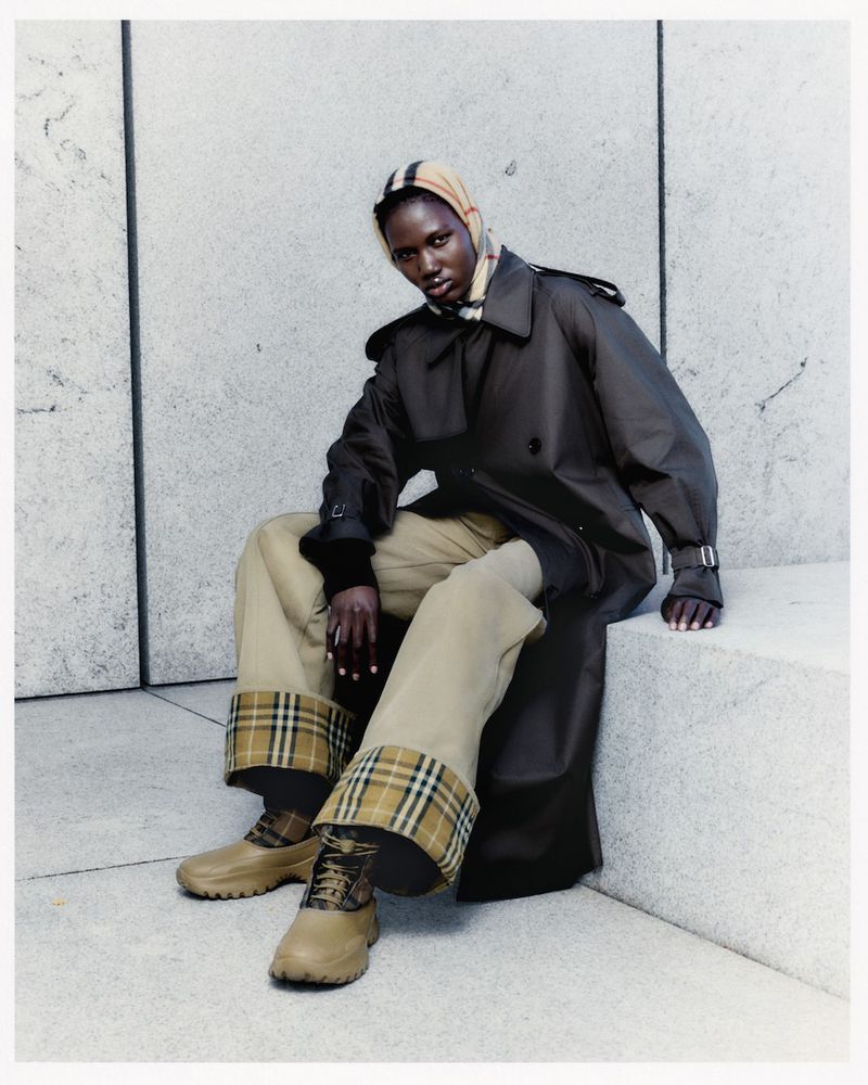 Woman in brown trench coat, beige pants, and matching shoes sits on a stone step, accessorized with a plaid headscarf and plaid detailing.