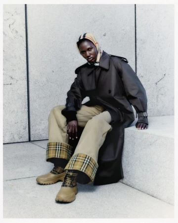 Woman in brown trench coat, beige pants, and matching shoes sits on a stone step, accessorized with a plaid headscarf and plaid detailing.