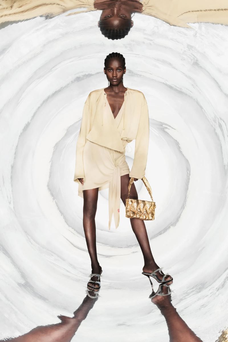 A lone fashion model poses in a cream-colored dress and high heels, holding a gold purse, in a reflective photo studio for a fashion editorial.