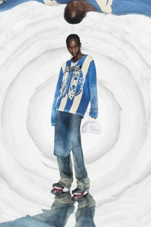 A slender model in a blue and white striped sweater and denim skirt, holding a white purse, stands against a swirling white background.