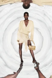 Model wearing a pale yellow draped outfit and holding a gold quilted handbag against a swirling circular abstract background.
