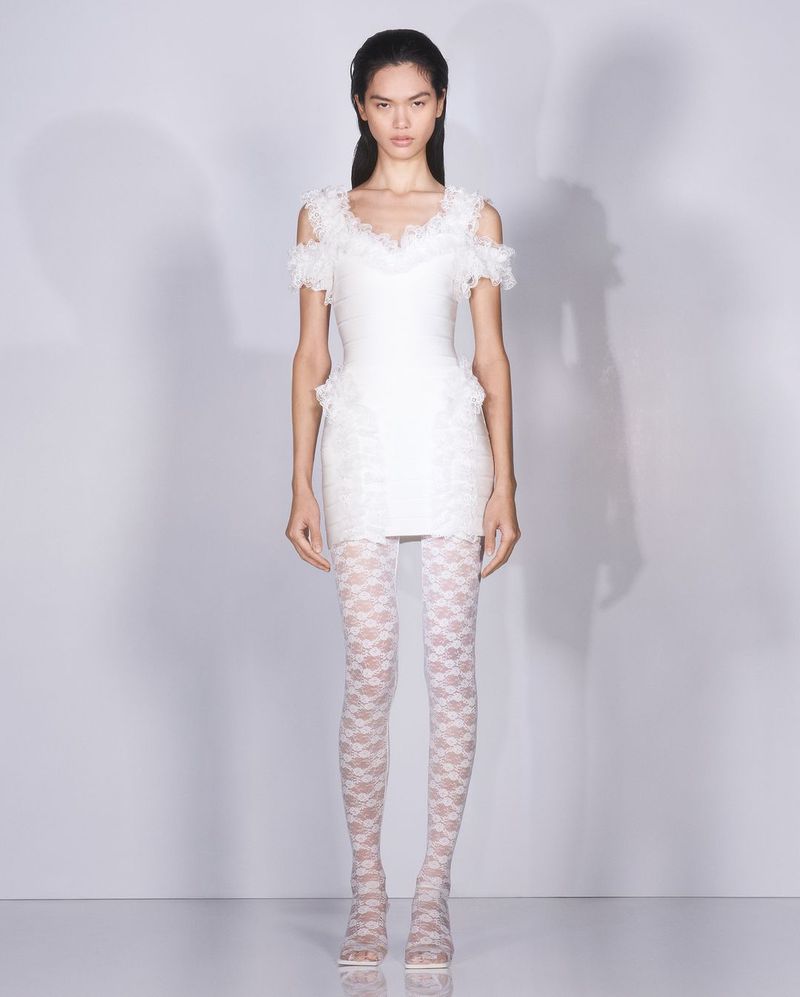 Full-length studio shot of a slender Asian woman wearing a short white lace dress and patterned white tights against a gradient white background.
