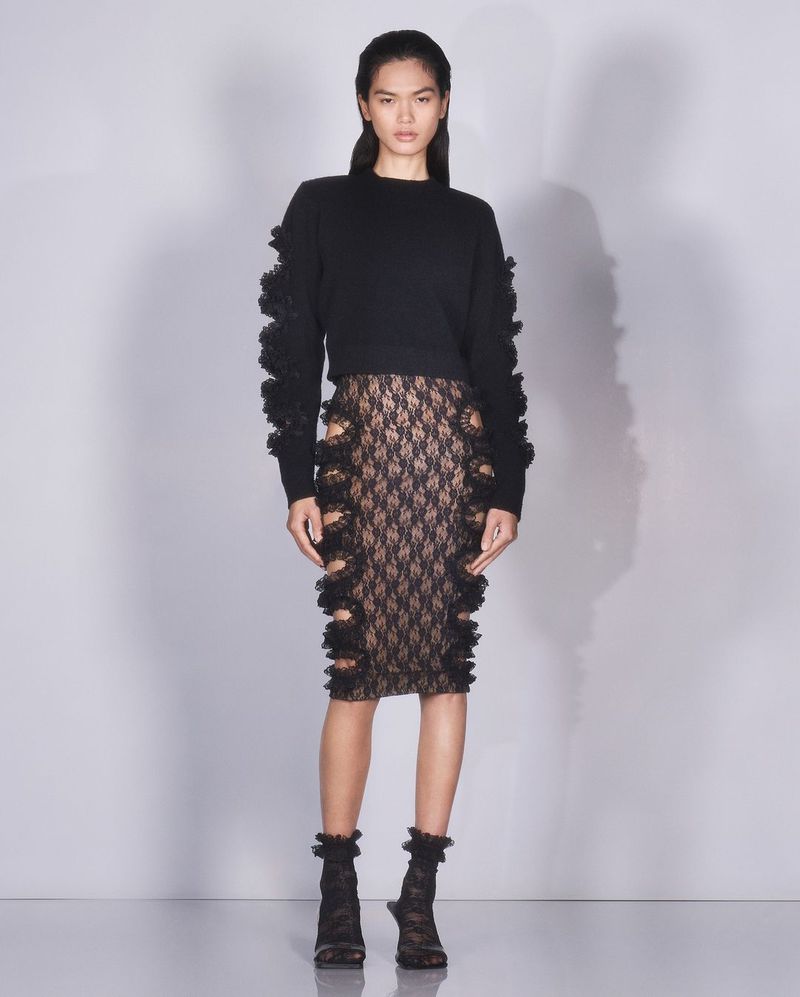 Elegant Asian woman in a black ruffled sweater and lace skirt poses in a bright studio, showcasing fashion and style.