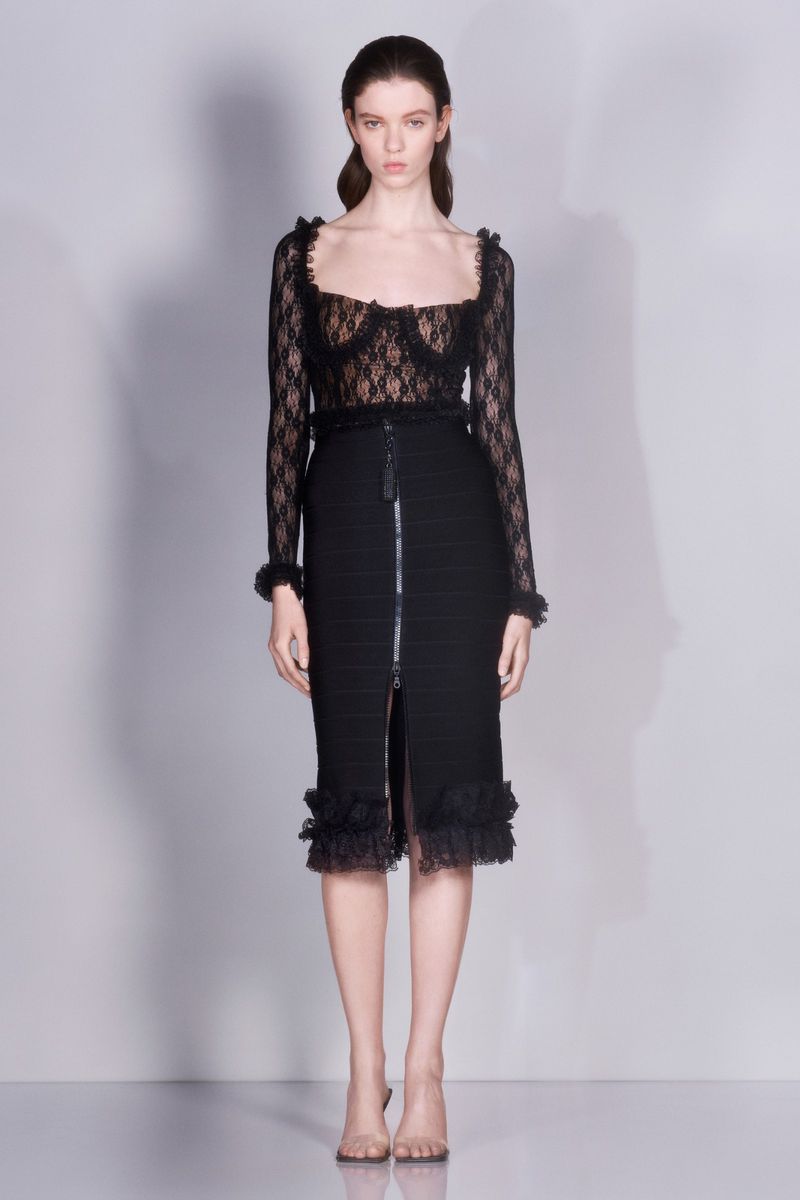 Full-length studio shot of a woman in a black lace long-sleeved top and pencil skirt with ruffles, posing against a light grey background.