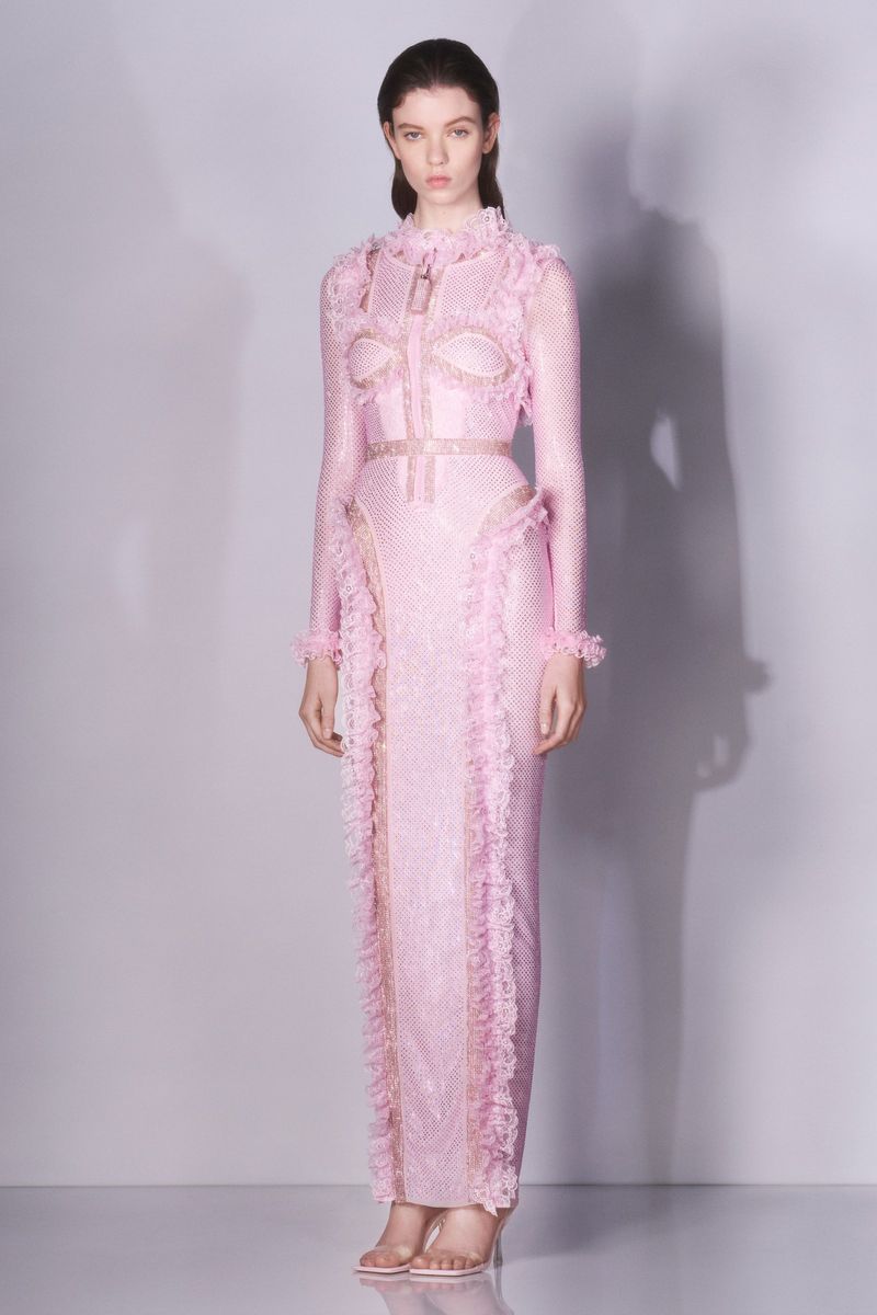 A woman poses in a long, pink gown with ruffled trim and long sleeves in a studio environment with a light gray backdrop.