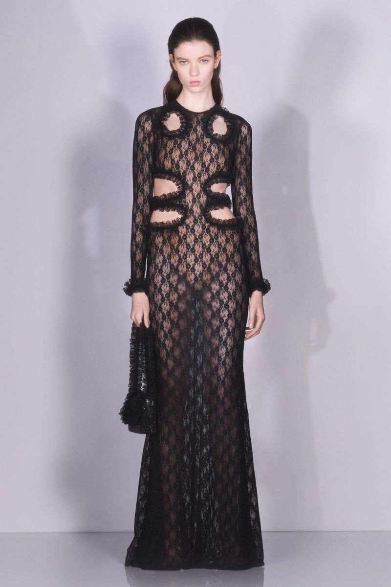 Elegant woman in a black lace dress stands in a photo studio with soft lighting and holding a small black purse. Fashion editorial.