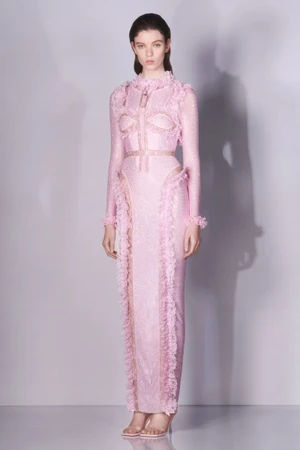 Model wearing a long pink textured dress with ruffles against a plain light grey studio background.