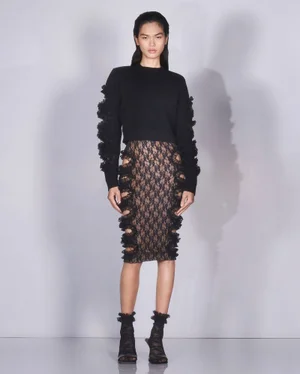 Model wearing a black sweater with textured ruffle sleeves and patterned lace skirt against a plain grey backdrop.