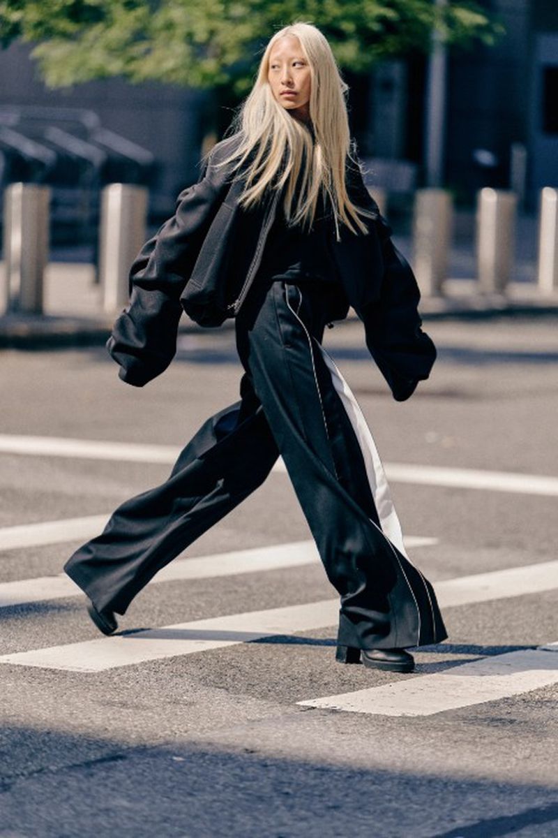 Asian woman with long blonde hair wearing a black jacket and baggy pants walks across a crosswalk in an urban environment on a sunny day.