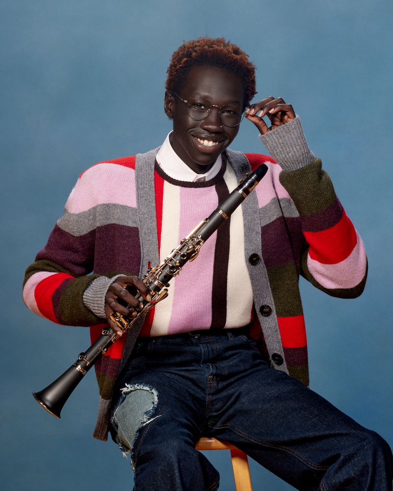Smiling African American man poses with a clarinet in a studio setting, wearing eyeglasses, colorful cardigan, and blue jeans.