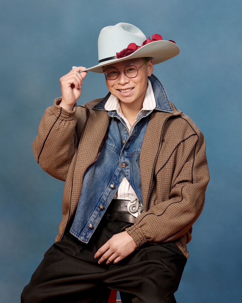 Stylish adult male in glasses and hat sits in front of blue backdrop wearing jacket over a denim shirt with a western belt buckle.