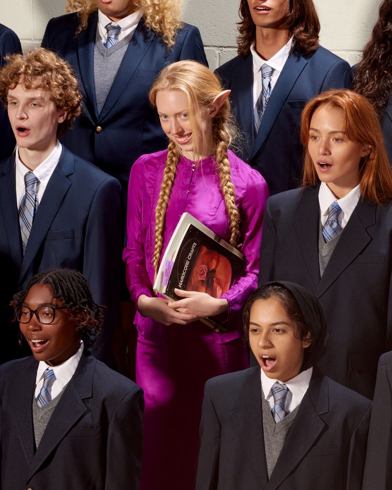 Group of young choir members in blue blazers, singing with open mouths. A woman in a purple dress stands out holding sheet music.