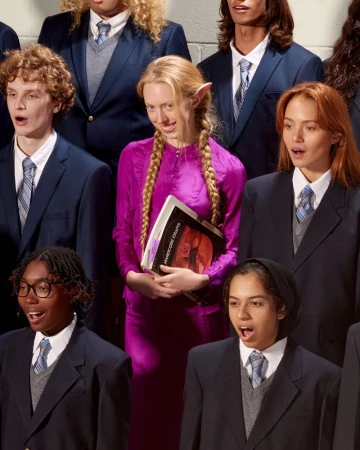 Group of young choir members in blue blazers, singing with open mouths. A woman in a purple dress stands out holding sheet music.