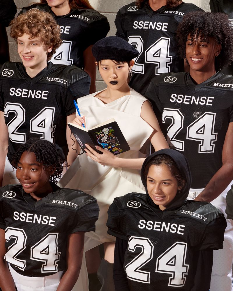 Diverse group in black football jerseys with 'SSENSE' and '24' posing with a person in off-white dress taking notes indoors.