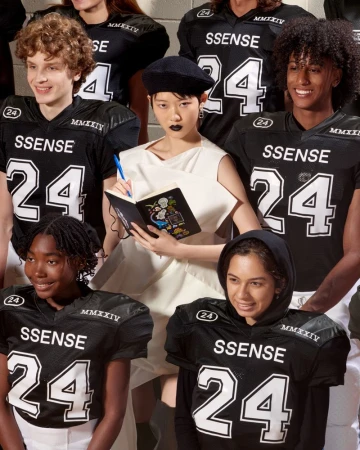 Diverse group in black football jerseys with 'SSENSE' and '24' posing with a person in off-white dress taking notes indoors.