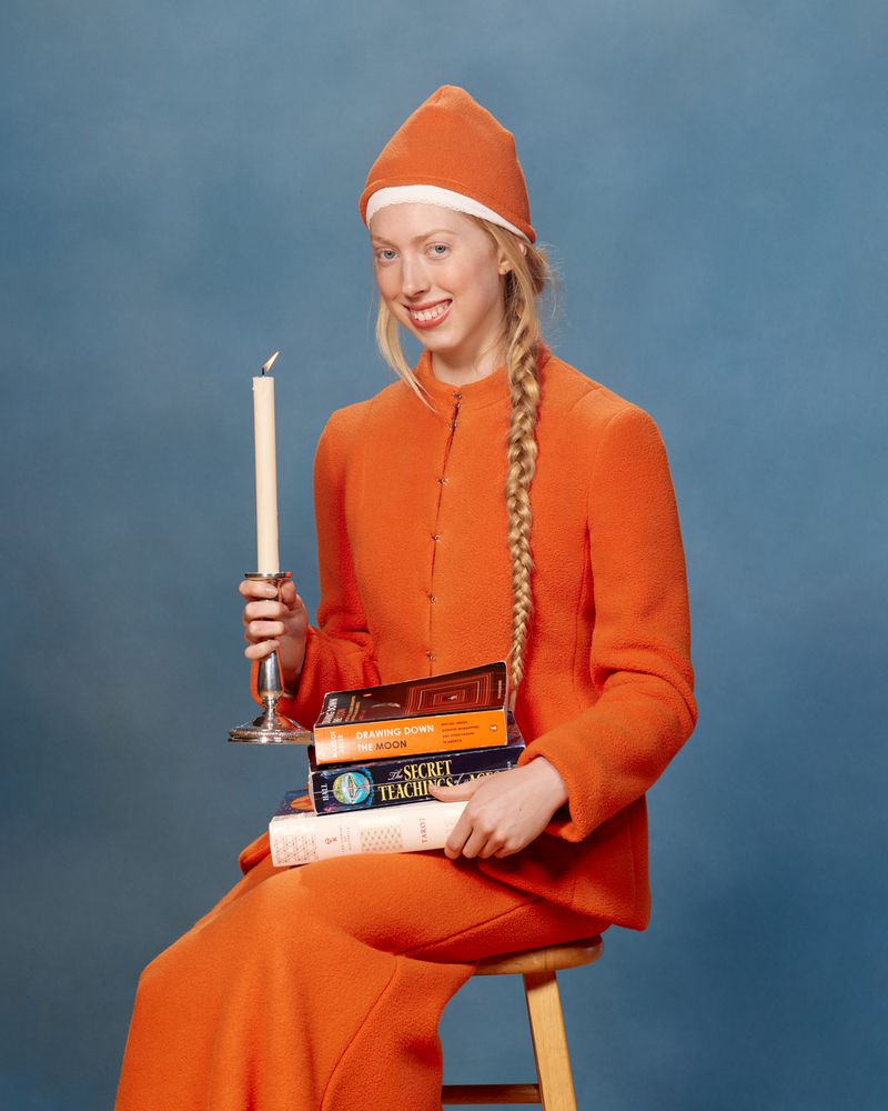 Blonde woman with a braided hairstyle wears an orange outfit and hat while seated, holding a candle and stack of books in a blue photo studio.